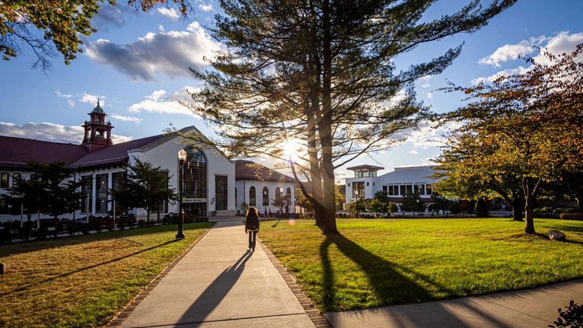 A person walks along a sunlit path on a picturesque campus with historic buildings and lush trees.