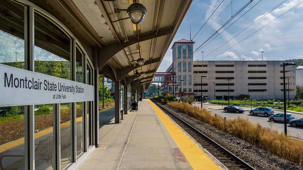 The image shows the platform of Montclair State University Station with a sign visible and a parking structure in the background.