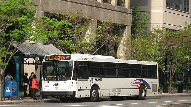 A NJ Transit bus is stopped at a bus stop with several people waiting nearby in an urban setting.