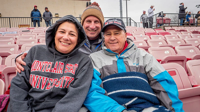 Three people, bundled up against the cold, are sitting in a stadium's bleachers, smiling at the camera.