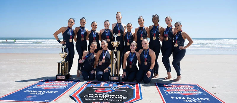 A cheerleading team poses with trophies and banners on a beach, celebrating their championship win.
