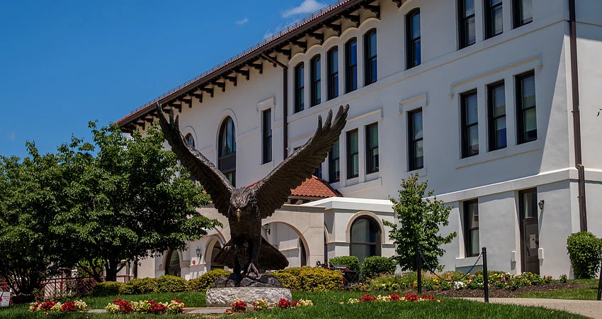 The image shows a large eagle statue in front of a white building with arched windows, set against a bright blue sky.