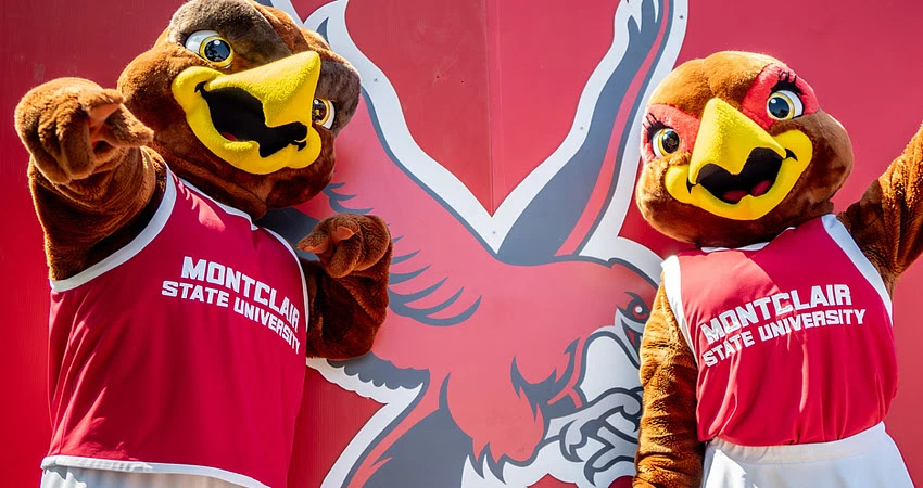 Two mascots in bird costumes, wearing Montclair State University jerseys, pose cheerfully in front of a matching backdrop.