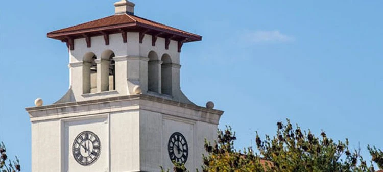 A clock tower with a red-tiled roof rises above the trees under a clear blue sky.