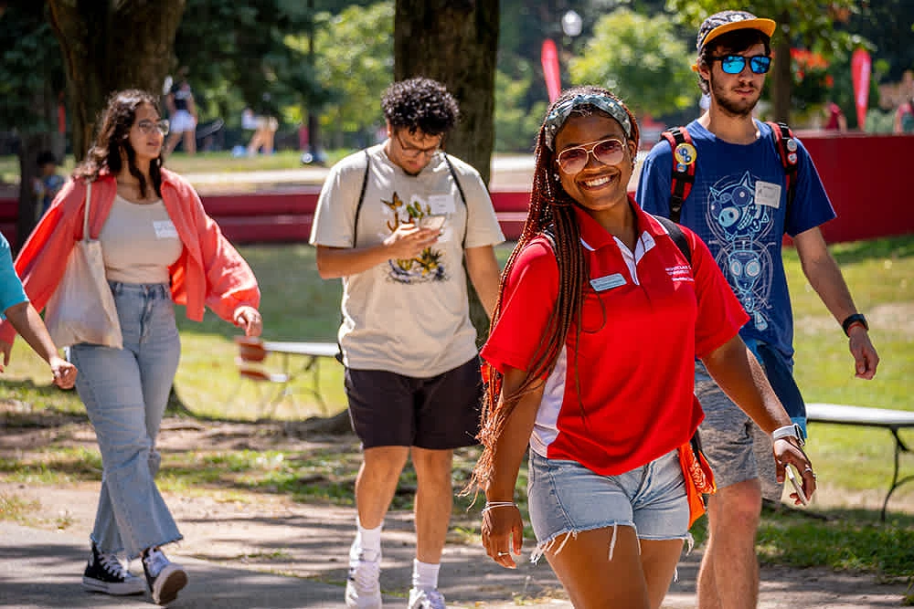 A group of college students, led by a person in a red shirt, walks outdoors on a sunny day.