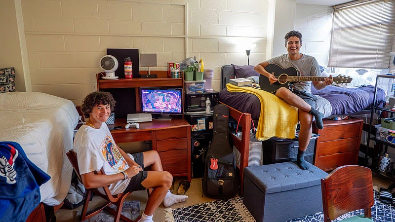 Two young men are enjoying their time in a dorm room, with one playing a guitar and the other sitting by a desk with a video game screen.