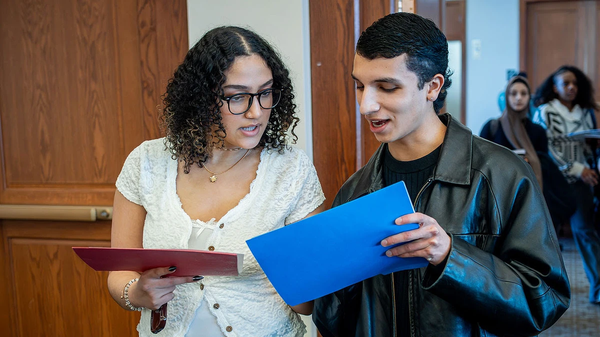 Two people are discussing something while holding folders, with others in the background.