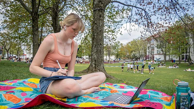 Student sitting on a blanket in the quad with iPad and Laptop.
