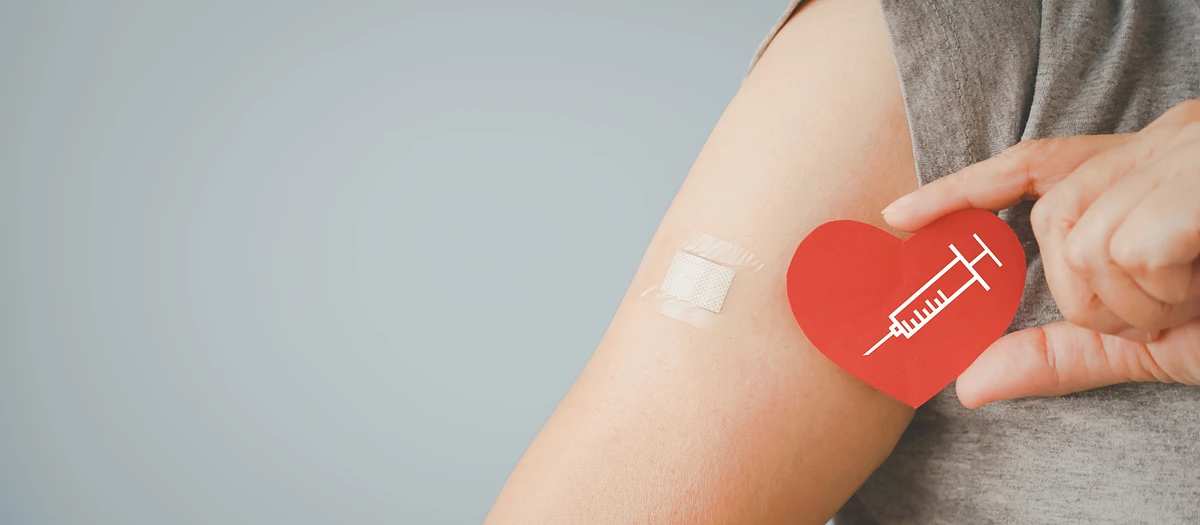 A person with a bandage on their upper arm holds a red paper heart featuring a syringe graphic, symbolizing vaccination.