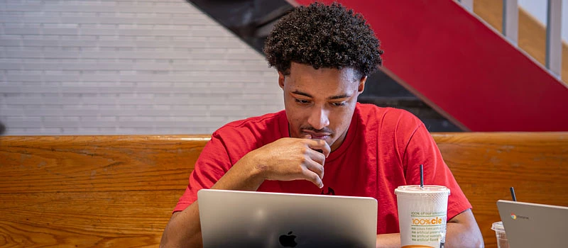 A person in a red shirt is intently working on a laptop at a table with drinks nearby.