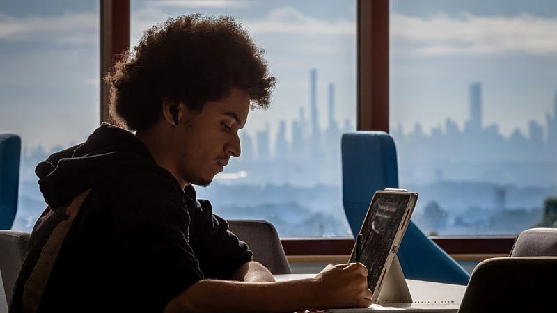 A person with an afro hairstyle is using a tablet while seated indoors, with a city skyline visible through the windows in the background.