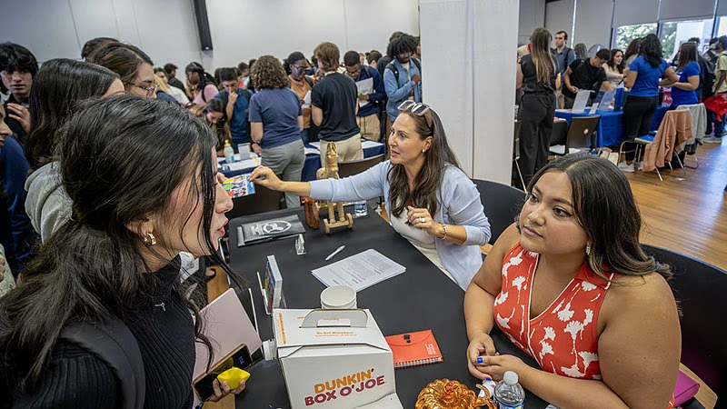 People are gathered in a busy room, with some having conversations at tables and others mingling or waiting in line.